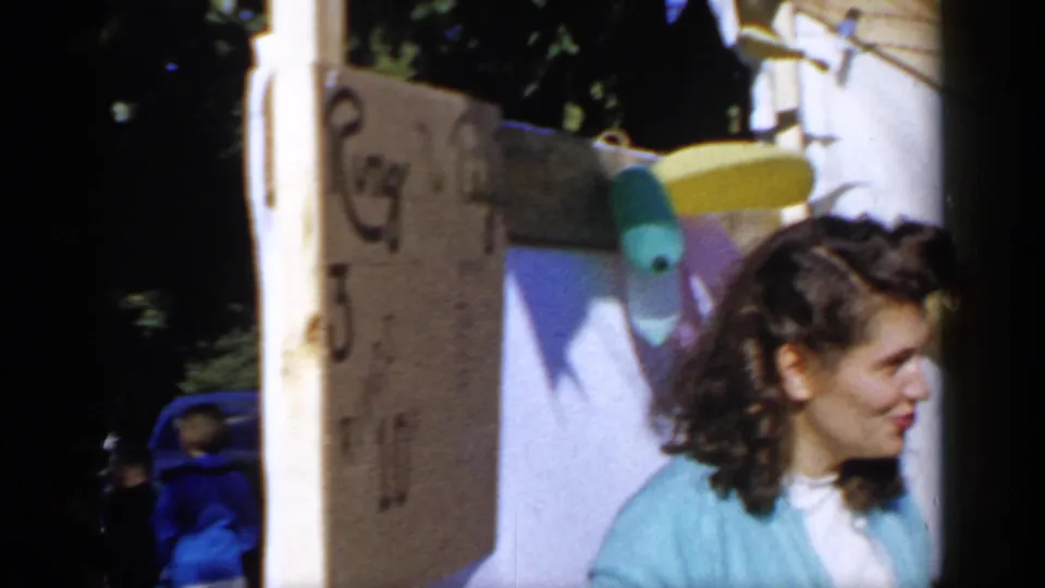 A woman in blue appears near a wall and a handmade-looking sign in the 1966 Mineola backyard home-movie scene.