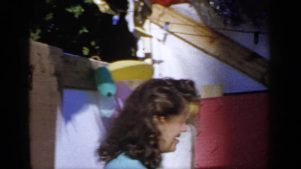 In 1966 Mineola home-movie footage, a woman in a light blue top smiles beside colorful outdoor decorations and wood trim.