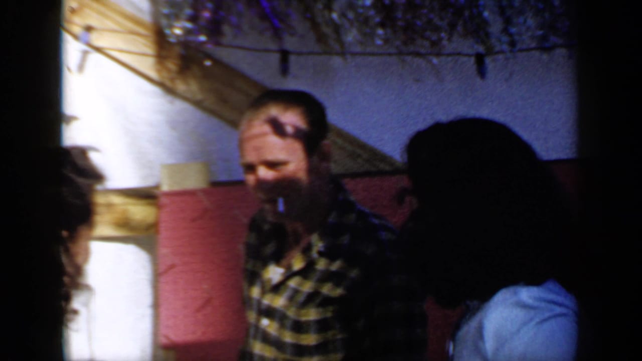 Man in a yellow plaid shirt stands near other adults beneath string lights.