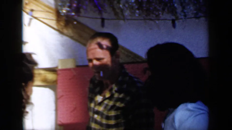 A man in a yellow and black plaid shirt stands among adults at the 1966 Mineola outdoor gathering, with string lights above.