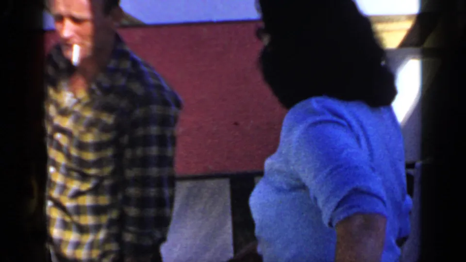 A man holding a cigarette stands near a woman in a blue shirt during the 1966 Mineola home-movie banquet scene.