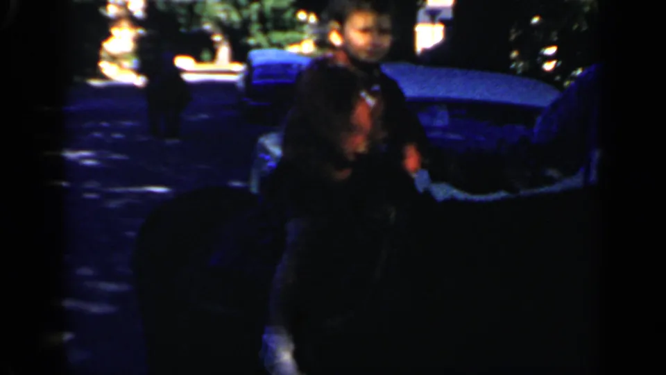 1966 Mineola home-movie frame showing people near parked cars and an outdoor gathering area, with heavy shadow and film-edge obstruction.