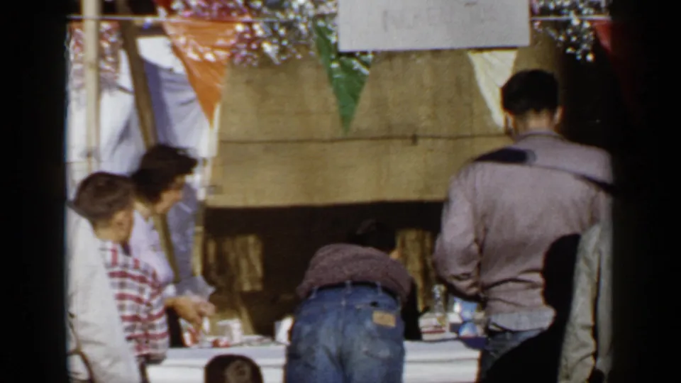 1966 Mineola event scene with adults and a child at a table beneath bunting, filmed from a casual home-movie viewpoint.