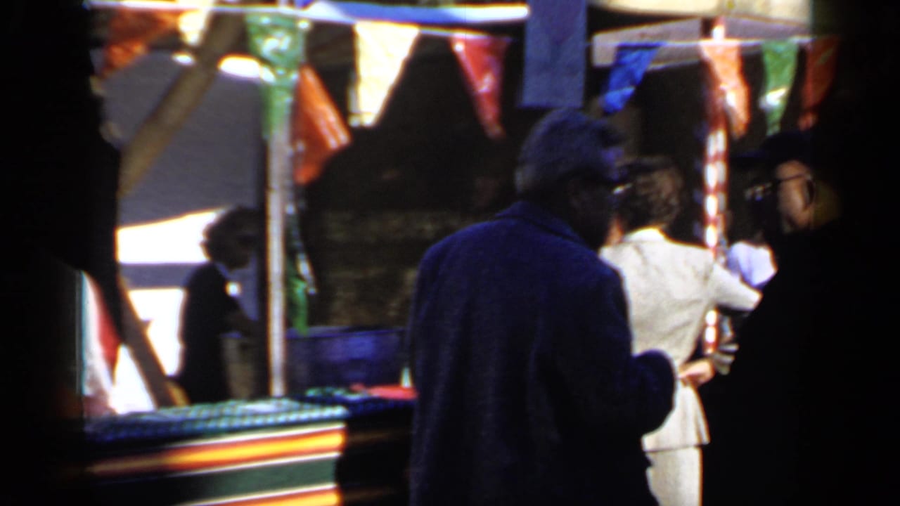 Adults stand near a striped booth under colorful bunting at a crowded outdoor event.