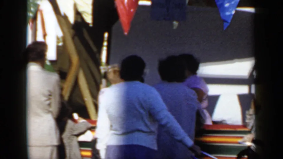 1966 Mineola home-movie frame with adults in sweaters and coats near bunting and a striped booth.