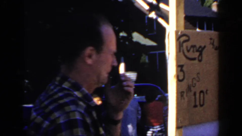 A man in a plaid shirt stands near a hand-lettered ring game sign advertising three rings for ten cents.