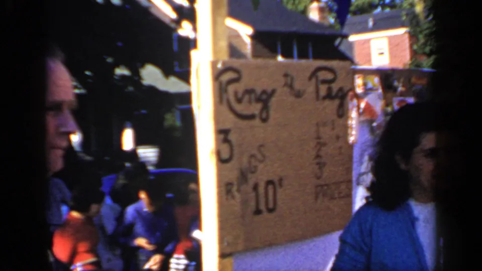 The ring-and-peg game sign and the woman in a blue cardigan share the frame at the Mineola carnival booth.