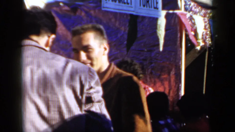 1966 Mineola home-movie frame showing young men in jackets near a street-side parade area with partial overhead signage.