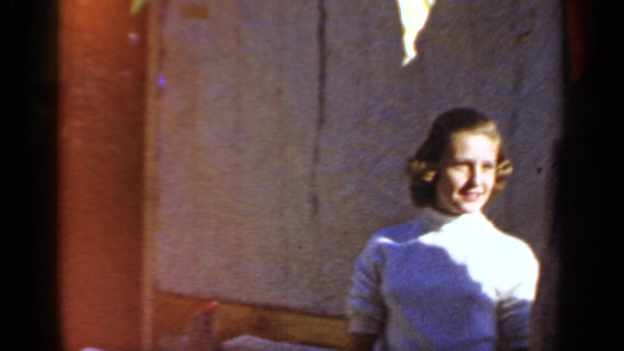 1966 Mineola home-movie frame showing a smiling woman in a white turtleneck near a sunlit wall.