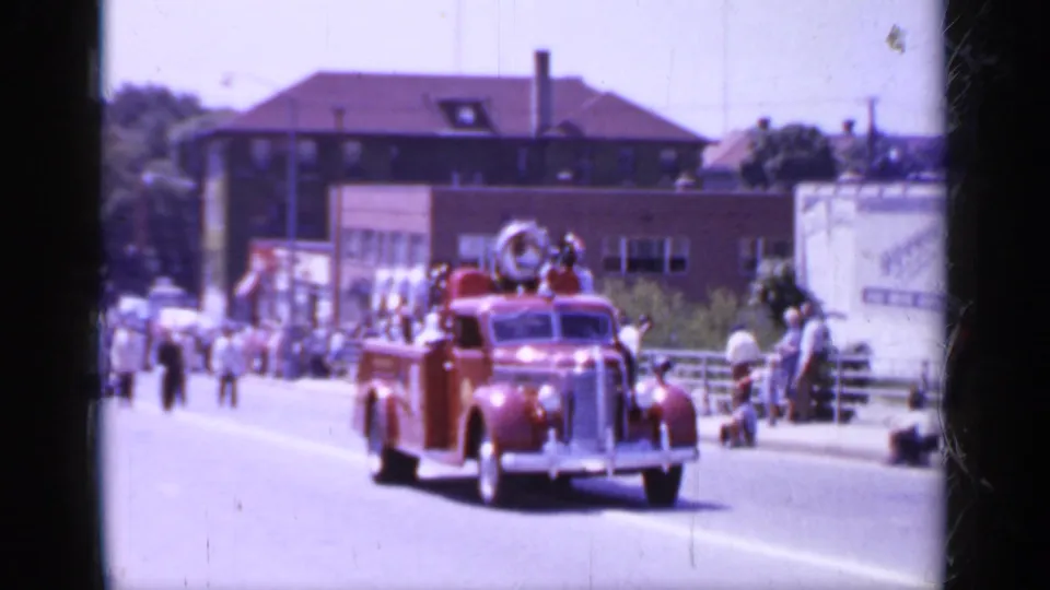 1966 Mineola parade frame showing a red firetruck approaching on a town street as spectators watch from both sides.