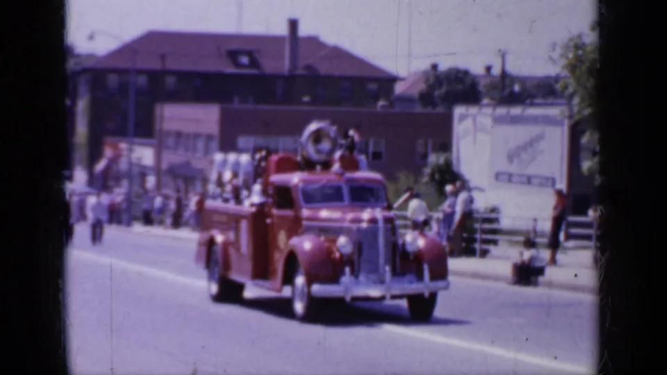 1966 Mineola home-movie frame showing a red parade firetruck moving past spectators and low town buildings.