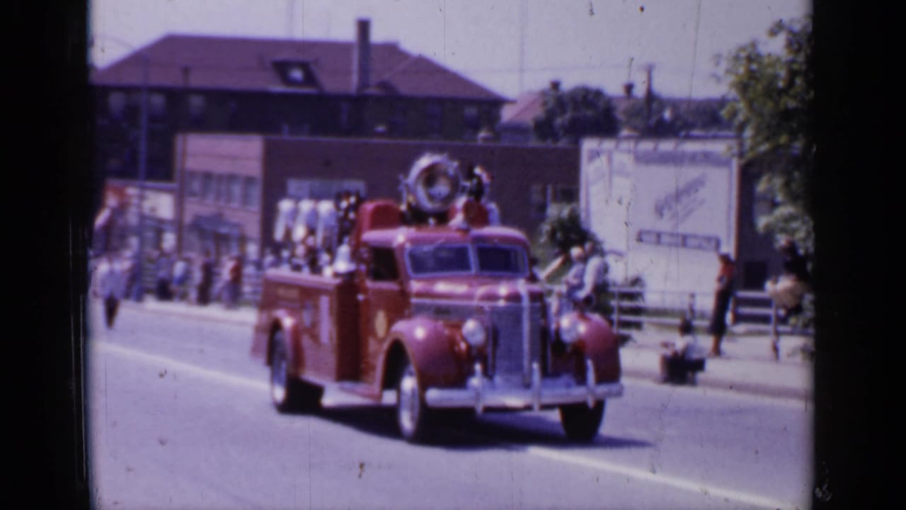 Red vintage fire truck approaches on a town street as spectators stand along the sidewalk.