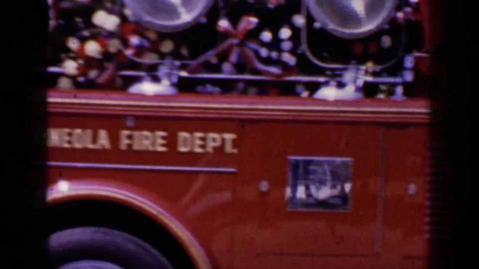 Close 8mm view of the red Mineola Fire Dept truck side, with lettering, lights, and equipment visible as it passes.