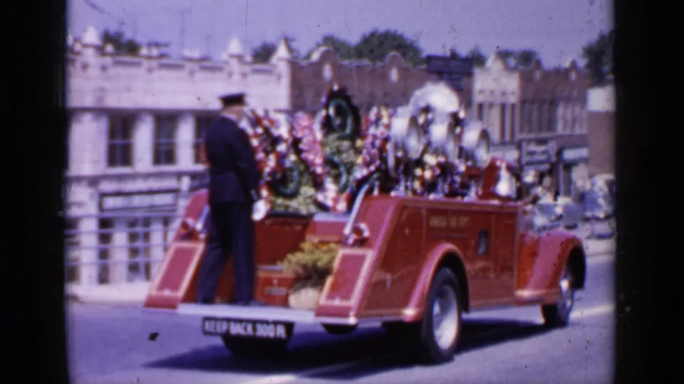 Rear 1966 Mineola view of the red fire department truck with floral wreaths and a uniformed firefighter standing at the back.