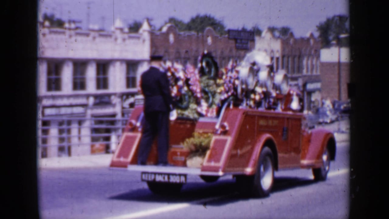 Decorated red fire truck moves along a town street with a uniformed man standing at the rear.