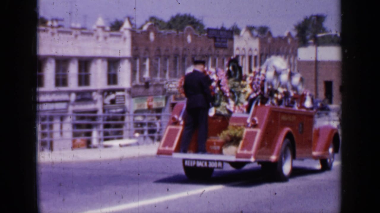 The decorated fire truck continues down a Mineola street, with floral displays, storefronts, and a rear warning sign visible.