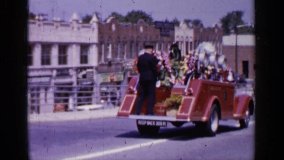 The decorated fire truck continues down a Mineola street, with floral displays, storefronts, and a rear warning sign visible.