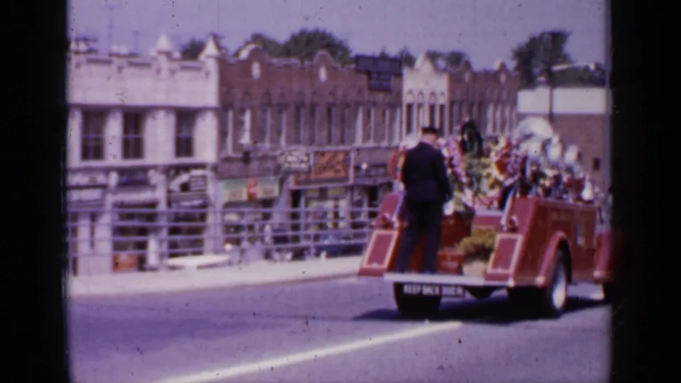 A soft 8mm frame shows the funeral procession vehicle moving away past Mineola storefronts and street railings.