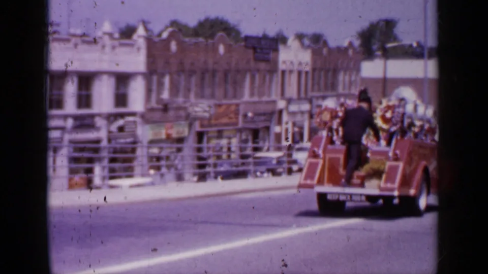 At the end of the Mineola procession shot, the decorated fire truck moves out of frame beside storefronts and railings.