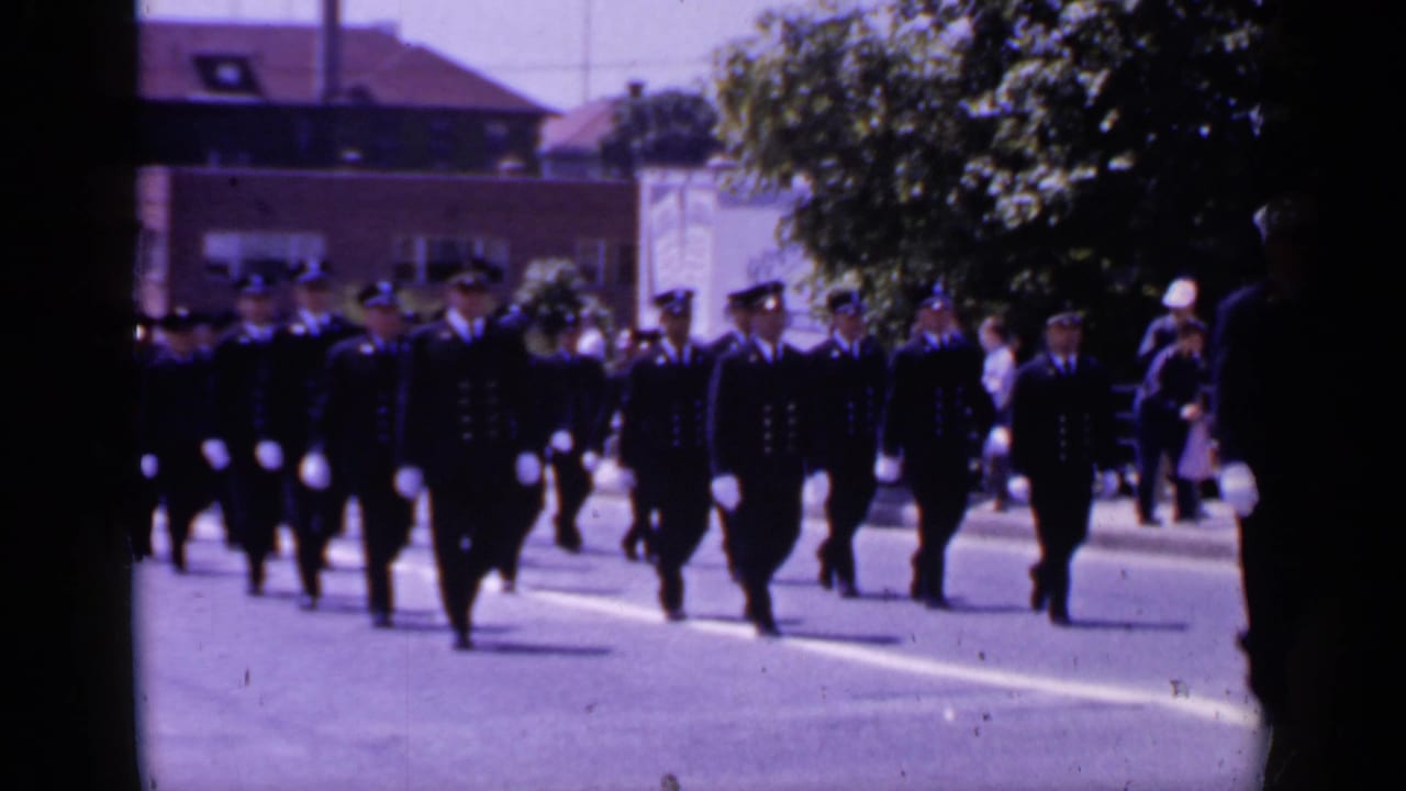 Uniformed men march in formation along a sunlit street.