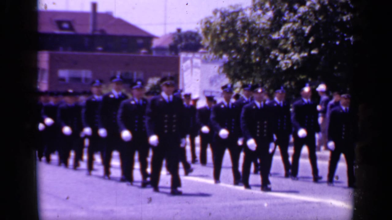 Uniformed men in dark parade dress move in synchronized rows along a Mineola street, with trees and brick buildings behind them.