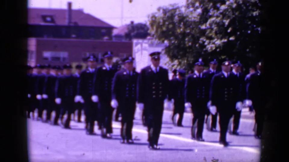 The 8mm frame shows a closer view of uniformed men in caps and white gloves marching in a formal Mineola parade formation.