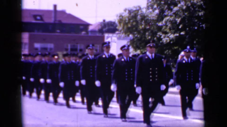 Men in dark uniforms, caps, and white gloves continue marching in formation through Mineola, with spectators and trees visible beyond them.