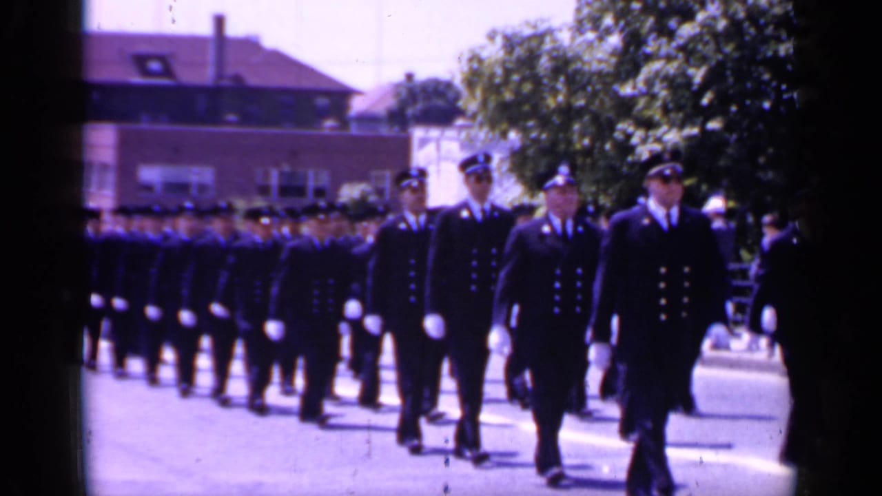 Uniformed cadets march in formation along a sunny street.