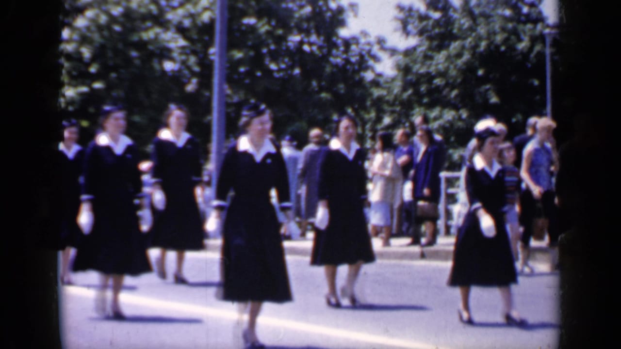 Uniformed women in dark dresses and white gloves walk along a sunny street.