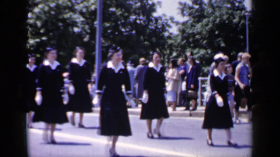 In Mineola, New York, 1966, uniformed women in dark dresses with white collars and gloves walk past sidewalk spectators.