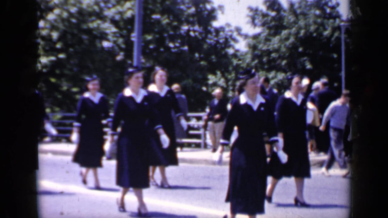 Uniformed women in dark dresses and white gloves pass by on a Mineola street as onlookers gather near roadside railings.