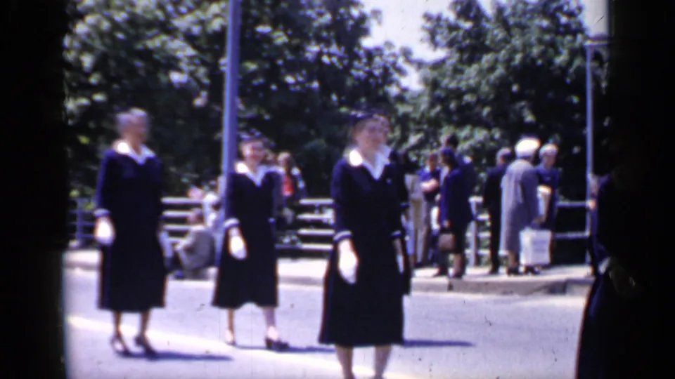 Two women in matching dark uniforms and white gloves walk through the foreground of a 1966 Mineola street scene.