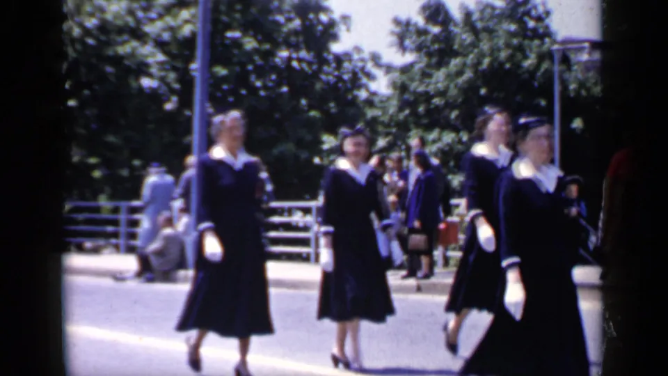 Women in dark uniforms with white collars and gloves continue along the sunlit Mineola street while bystanders look on.