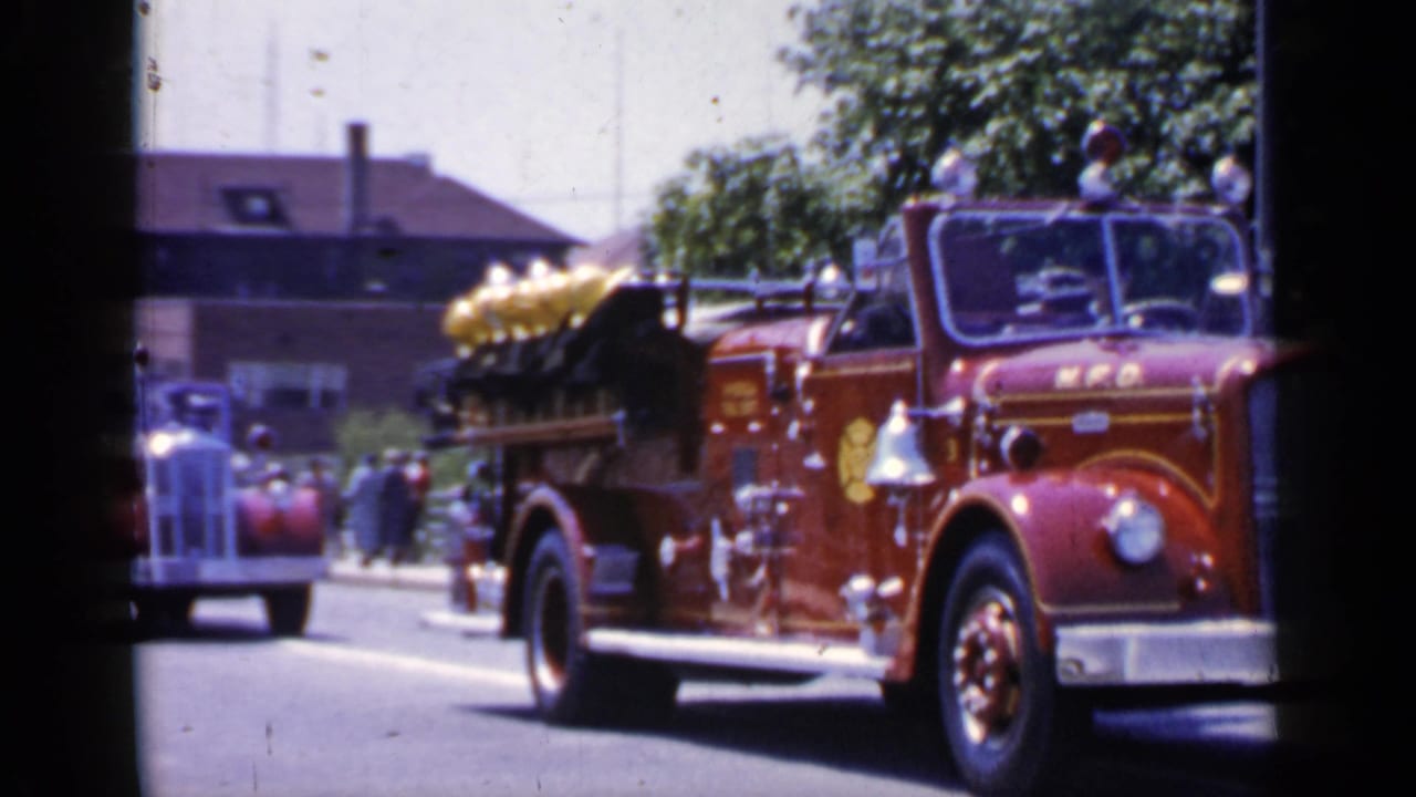 Red fire engine passing a vintage red car on a sunlit street with spectators nearby.