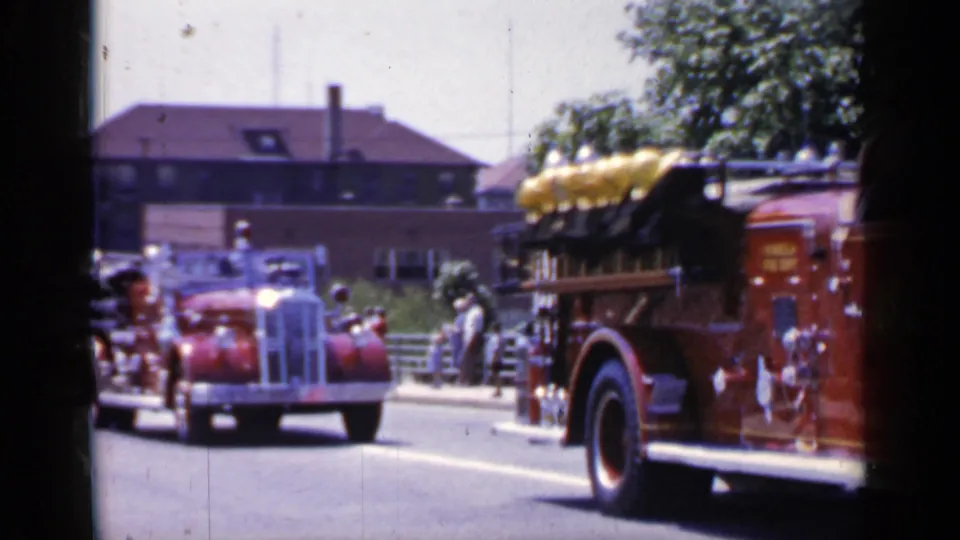 1966 Mineola, New York frame showing a vintage red fire car approaching behind a passing fire engine during the parade.