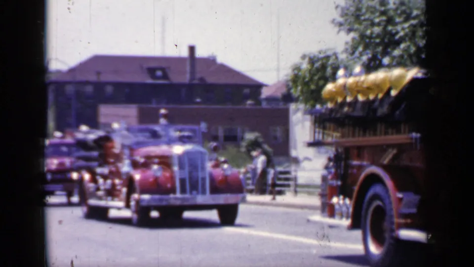 1966 Mineola parade frame with a red vintage car centered behind a fire engine, viewed from a handheld curbside position.