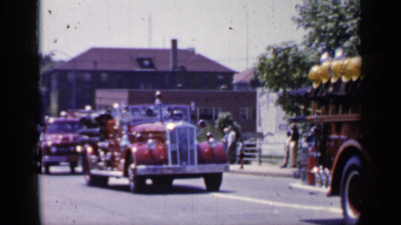Red antique fire engine leads a street parade with another red vehicle behind it.