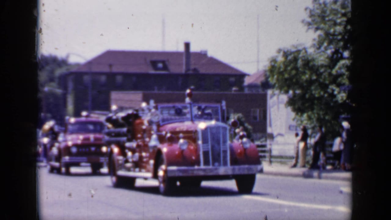 Red antique fire engine drives toward the camera on a sunny street.