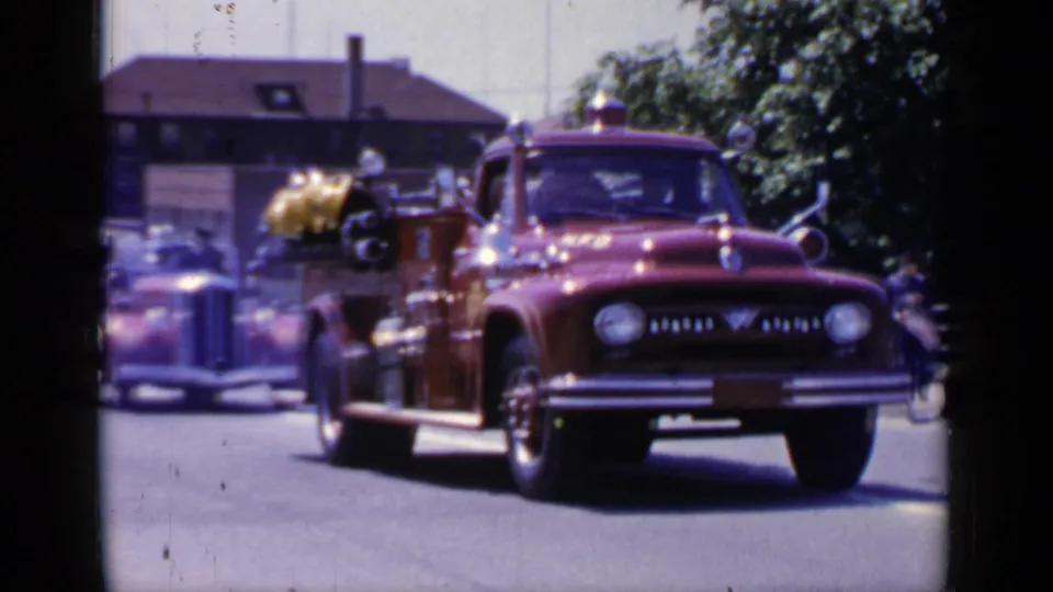 A red rescue truck, likely a mid-century Ford-style vehicle, follows an older fire engine in the Mineola, New York parade footage.