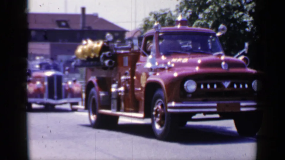 1966 Mineola home-movie frame showing a red fire truck passing spectators on a sunlit street.
