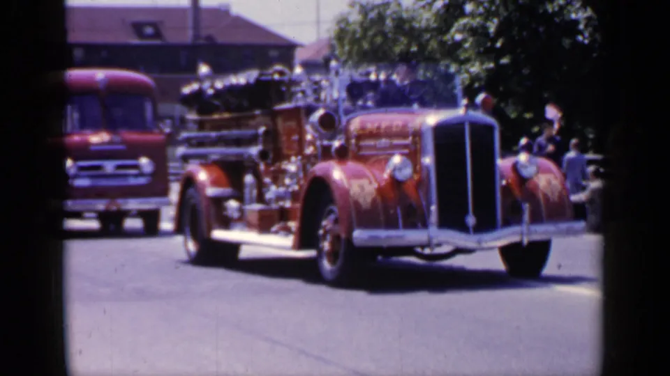 1966 Mineola home-movie frame showing an antique red fire engine rolling past a curbside crowd.