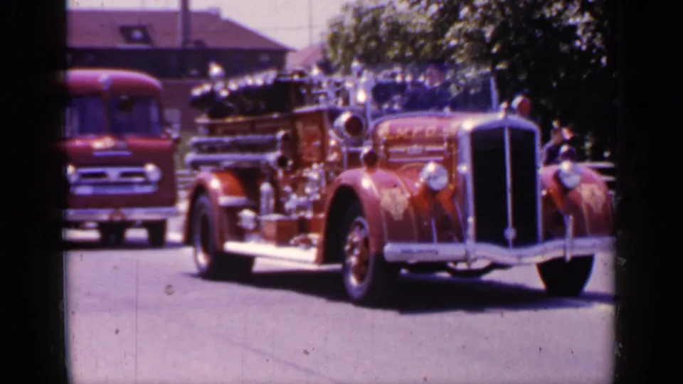 1966 Mineola frame with a red antique fire engine near the camera and spectators along the route.