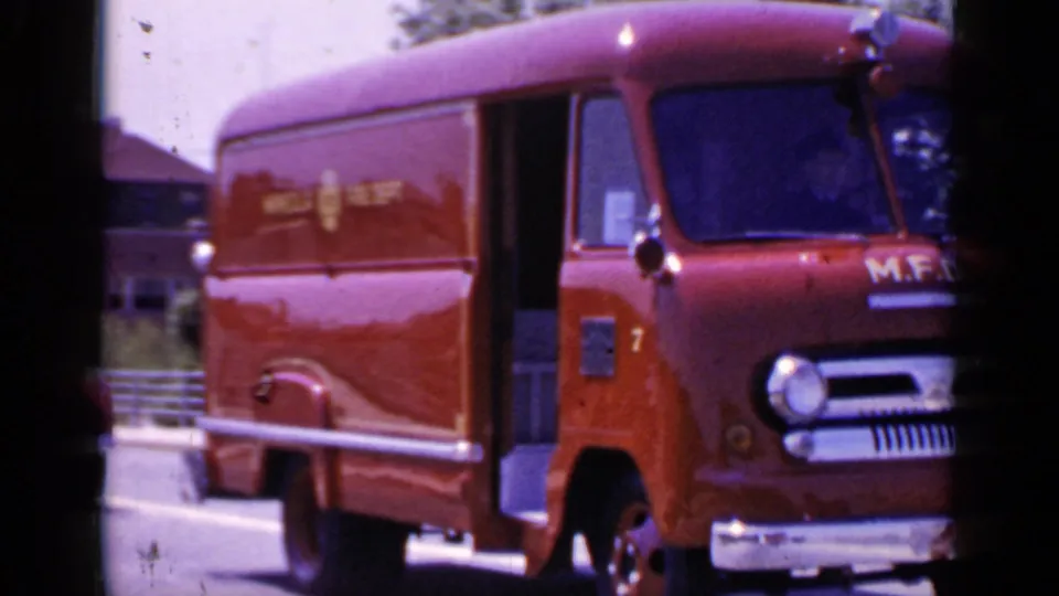 A red Mineola Fire Dept. van moves by on the street, showing side lettering and a rounded 1960s body shape.