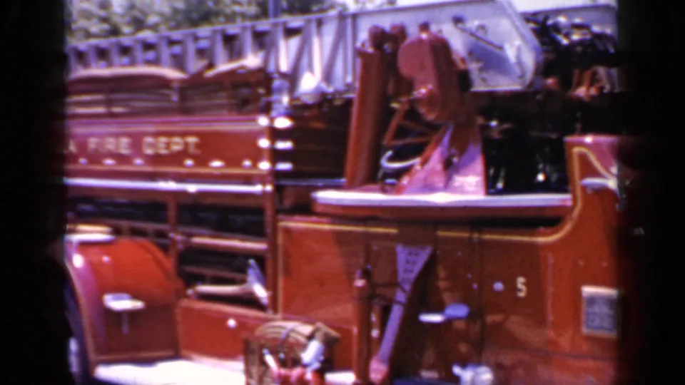 Close 8mm view of the Mineola Fire Department truck side, showing the open cab, mounted bell, ladders, and painted department lettering.