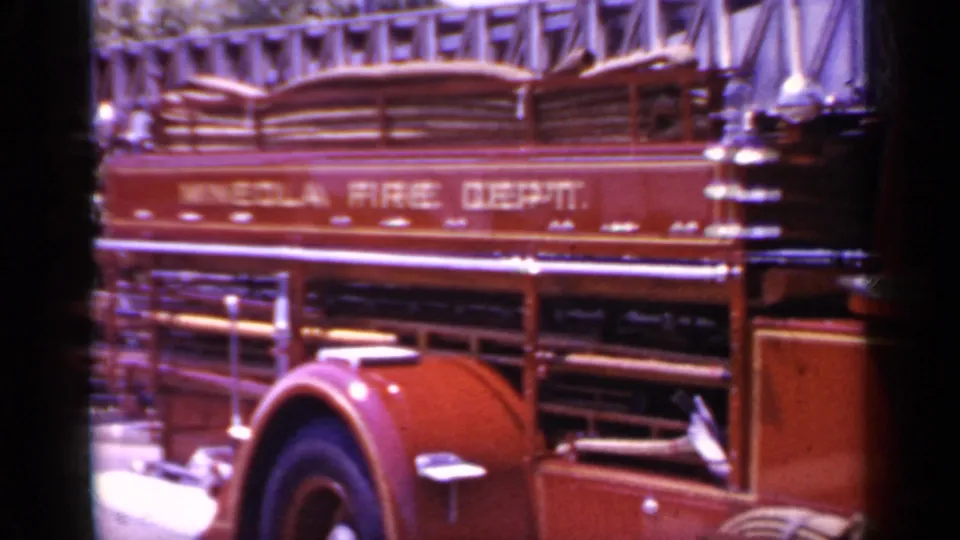 1966 8mm frame of the Mineola Fire Dept. truck side, emphasizing the painted department name, ladders, wheel arch, and polished hardware.