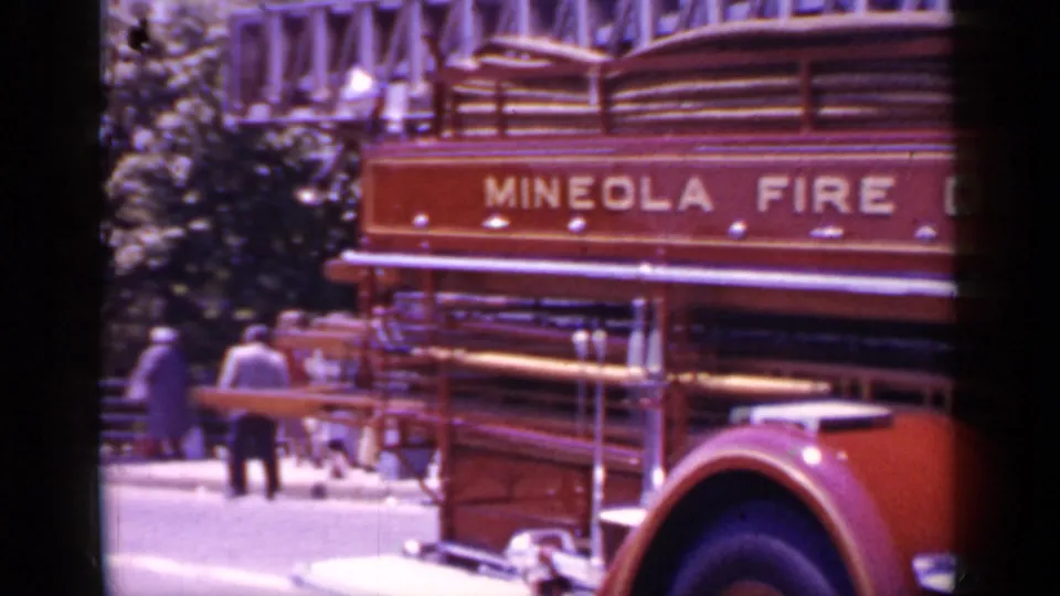 1966 Mineola, New York frame showing the rear side of the fire truck with ladders, painted lettering, and distant bystanders outdoors.