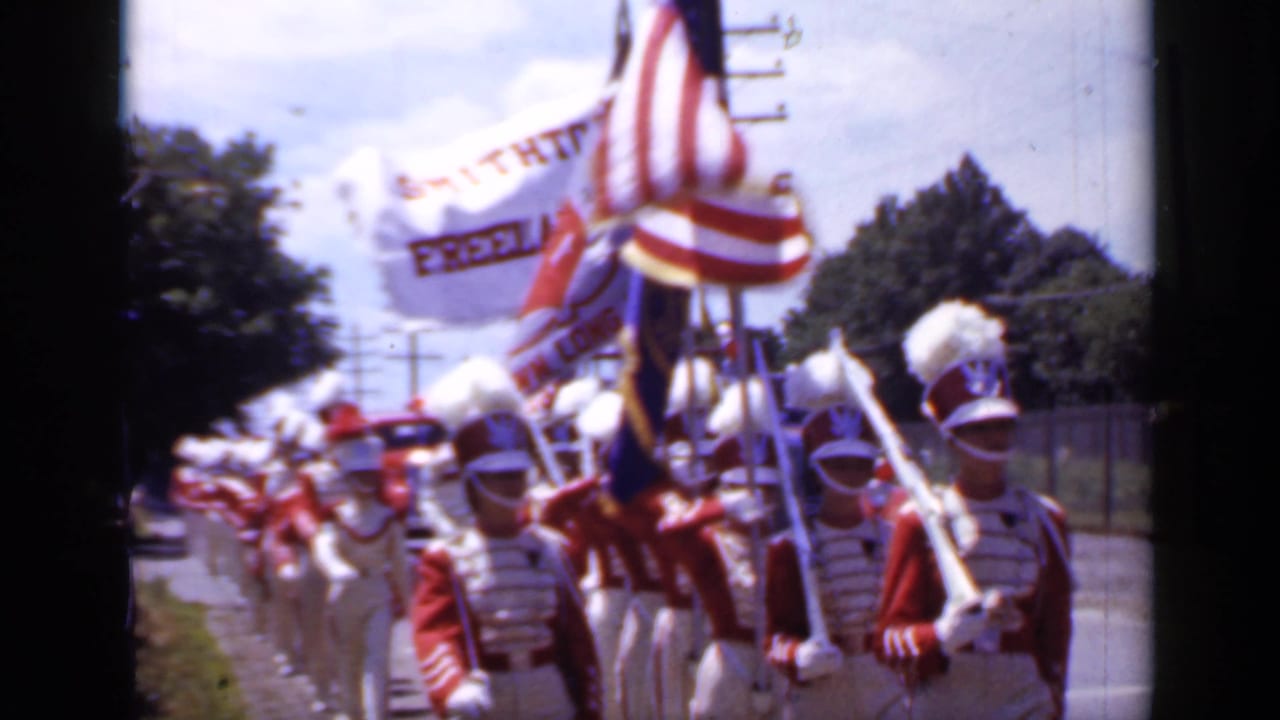 Marching band in red and white uniforms carrying an American flag along a suburban street.