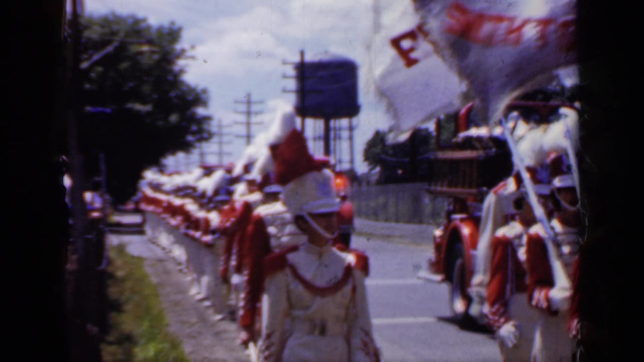 Parade marchers in red and white uniforms move down the road near a water tower.
