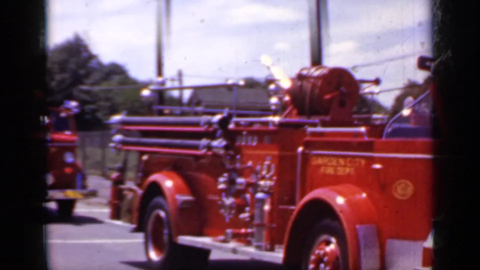 A red Garden City Fire Department truck moves through the Mineola parade route, filmed from a close curbside home-movie angle.