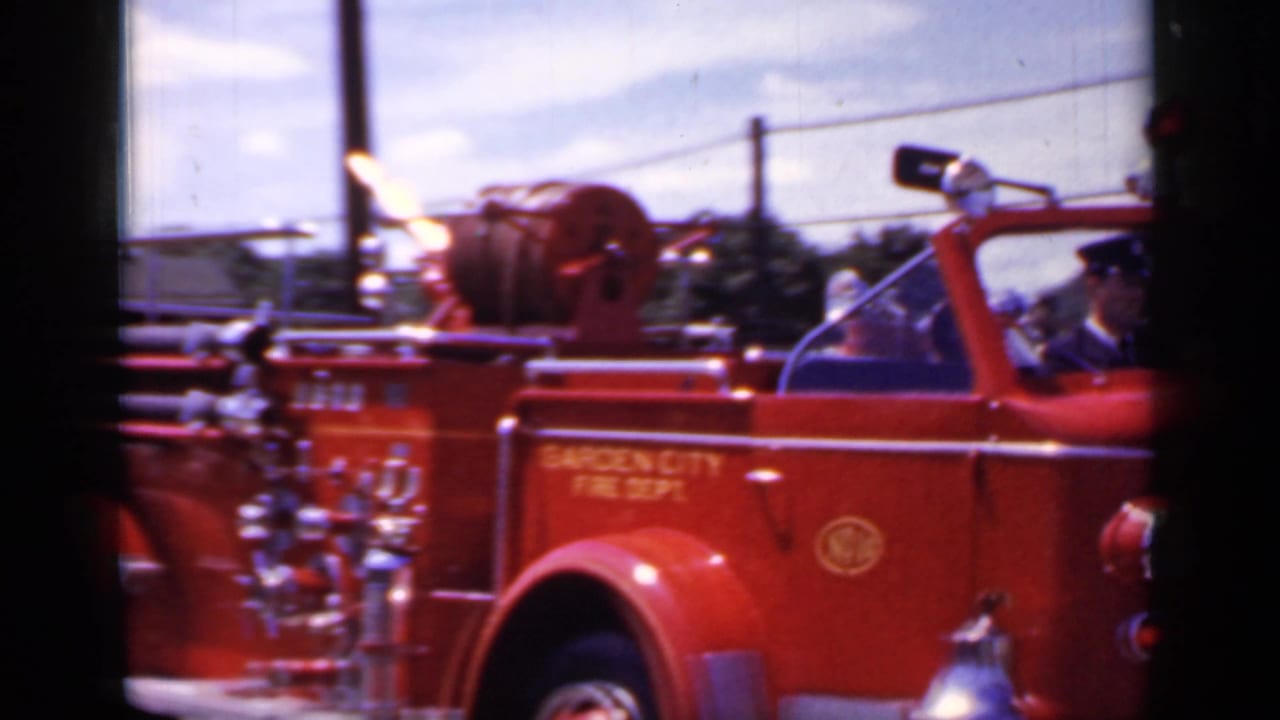 Red open-cab fire truck passing on a sunny street, with utility poles behind it.